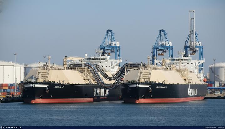 Два танкера-газовоза стоят борт о борт в промышленном порту и соединены шлангами для перевалки груза.