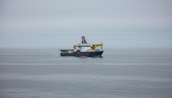 Морское исследовательское судно в открытом море. На корме виден кран для спуска подводного оборудования. Спокойная вода, пасмурная погода.